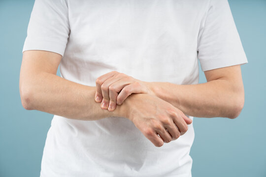 Close up of male hands on blue studio background holding wrists, suffering of pain or discomfort. Gesture suggests physical strain, injury or repetitive stress. Carpal tunnel, inflammation, arthritis