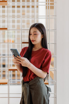 Young asian woman wearing a red t-shirt using a smartphone, browsing social media, chatting, or shopping online, connecting with technology and digital communication indoors