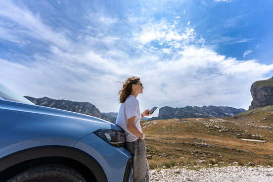Woman with smartphone taking a break by her blue car during a mountain travel journey.