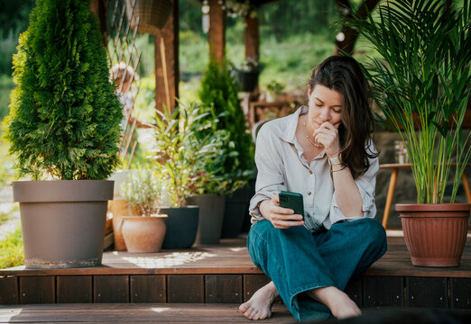 Pensive summer mood. Thoughtful young woman holding a cellphone while sitting on a wooden cottage patio.