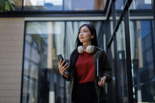 Young asian woman carrying laptop and smartphone, wearing headphones around neck, walking in modern city during daylight, looking confident and focused