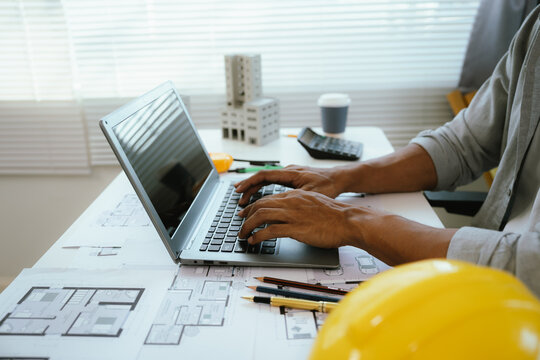 An architect or engineer works on a laptop with building plans and safety equipment, representing construction planning, project management, architecture, and engineering design.