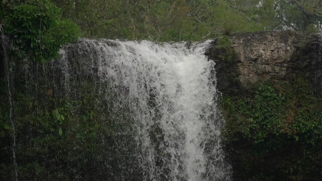 Static slow motion shot of the top section of Tad Champee waterfall, showing water spilling over the rocky ledge and surrounding vegetation.