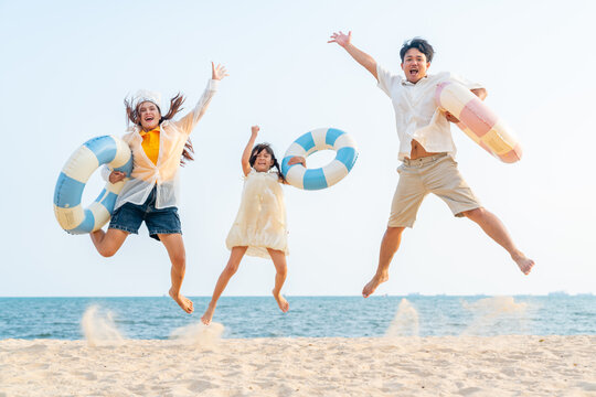 Portrait of Happy Asian family parents and child holding swim ring and jumping at beach. Father, mother and little daughter enjoy outdoor active lifestyle travel the ocean on summer holiday vacation.