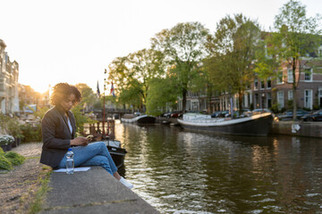 Fototapeta premium Woman freelancer using smartphone while sitting on a canal wall in amsterdam during golden hour
