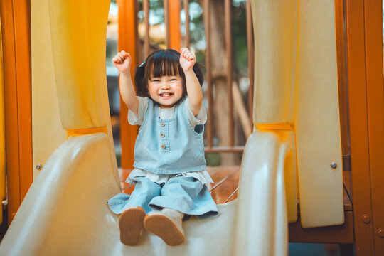 Cheerful Asian toddler girl raising her arms in excitement while sliding down a yellow slide at an outdoor playground. Pure childhood joy, Family weekend activities. Parenting, child development