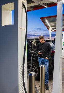 Man using phone while charging his electric SUV at dusk