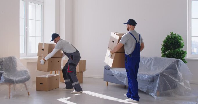 House removals and delivery company workers deliver boxes in new apartment. Two young men come in living room interior with furniture in plastic covers, put boxes on floor and then go to bring more