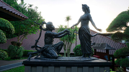 Sunset glows behind bronze statues of two figures. One kneels, bowing low in respect or worship. The other stands tall, arms extended gracefully. They appear serene, as if in quiet contemplation © Cak Had Photography