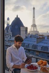 Obraz premium Breakfast, Eiffel Tower and tablet with man on balcony of hotel for holiday, travel or vacation. App, food and landmark with tourist on terrace of accommodation for sightseeing in Paris, France