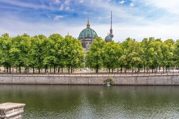 Berlin Cathedral and the Fernsehturm rise behind rows of green deciduous trees. The river and cloudy blue sky create a calm city panorama. © Jakob