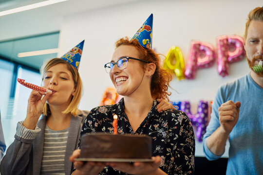 Colleagues having a birthday celebration in office with cake, party blower and party hats