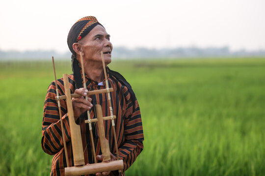 An elderly Indonesian man in a rural setting, wearing a traditional Javanese lurik surjan shirt and blangkon headpiece, proudly holds a bamboo angklung musical instrument.