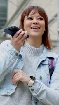 Vertical video: Looking down, woman raising phone, recording message outside building, denim jacket