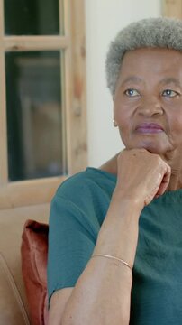 Vertical video: Holding chin, senior shifting gaze and thinking on sofa by wood window in teal top