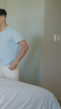 Vertical video: Entering from right, man in blue tee making bed with white sheet in bedroom