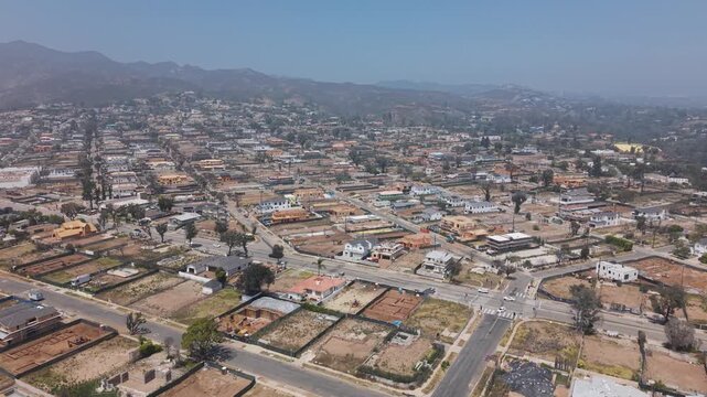 Pacific Palisades wildfire aftermath showing scattered empty lots and construction frames across residential grid, dry brown hills stretching under hazy sky. California, USA. Aerial wide shot.