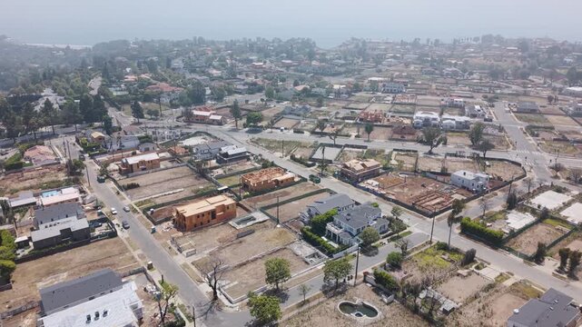 Pacific Palisades wildfire aftermath with cleared lots and reconstruction beside surviving homes, palm trees and hazy coastline in distance under pale sky. California, USA. Aerial flyover.