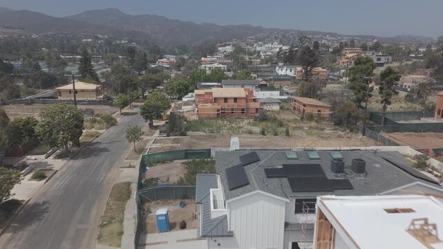 Pacific Palisades wildfire aftermath with construction frames on street corner, surviving homes and dry hills with scattered trees beyond under hazy blue sky. California, USA. Aerial flyover shot.