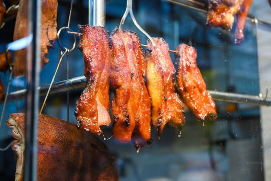 Close up of traditional Cantonese char siu honey roasted pork and crispy pork belly hanging on metal rack behind glass window of restaurant