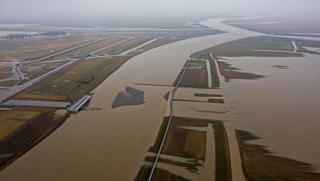 Aerial view of a wide brown river flowing through a flooded landscape with fields and dikes under a cloudy, overcast sky in a rural region