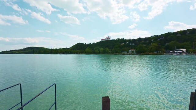 Scenic view across Lake Balaton toward the Tihany Peninsula and the historic Tihany Abbey overlooking the shoreline. Peaceful lakeside landscape with turquoise water, forested hills, and dramatic clou
