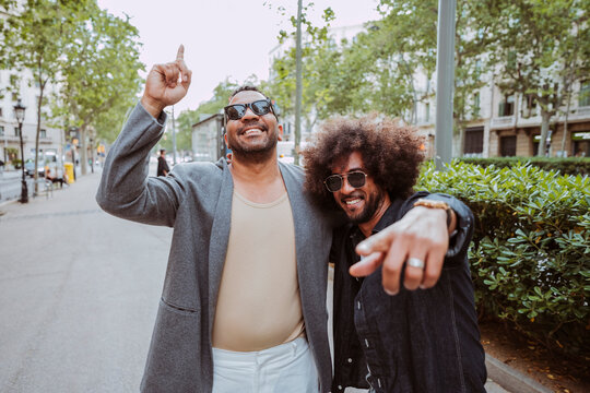 Two smiling latin american men wearing sunglasses enjoying camaraderie while pointing finger at camera on a city street.