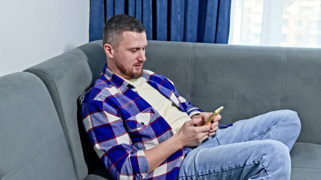 A man is sitting comfortably on a gray sofa in a living room. He is focused on his smartphone while the sunlight shines through the window.