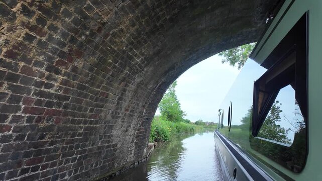Looking along the side of a green narrowboat cruising along a canal. The boat passes under a historic brick canal bridge, and there is beautiful and calming rural scenery all around.