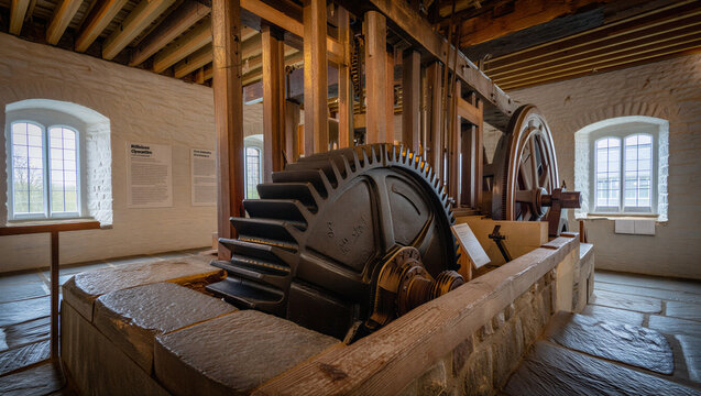 Massive cast iron gears and wooden support beams inside a historic windmill or watermill machinery room with stone walls and windows