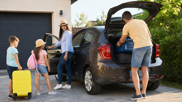 A family is getting ready to go on a trip. One parent is loading bags into the car while two kids are waiting near the vehicle. The scene takes place in a driveway on a sunny day.