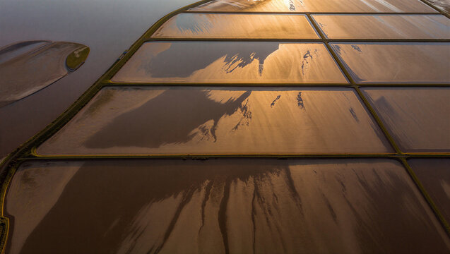 Aerial view of salt evaporation ponds with geometric shapes, brown water, and reflective surfaces under the warm light of a setting sun
