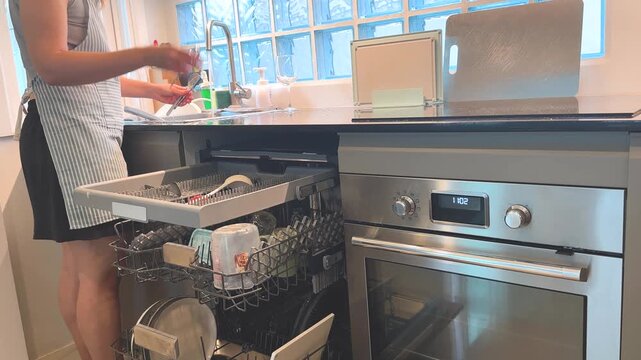 Woman rinsing dishes by hand and loading them into open dishwasher in home kitchen. Real life video footage of dishwashing, kitchen cleaning, household chores and everyday domestic routine.