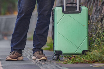 Man stands beside green suitcase on sidewalk. He wears jeans and sturdy boots. Luggage rests near grassy edge. Urban setting, casual travel moment. Ideal for travel, adventure, or transit themes © Anzz Stock