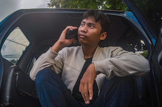 Young man sits in car, talking on phone. He looks thoughtful, focused on conversation. Light filters through trees, softening scene. Urban setting implied, perhaps waiting or pausing