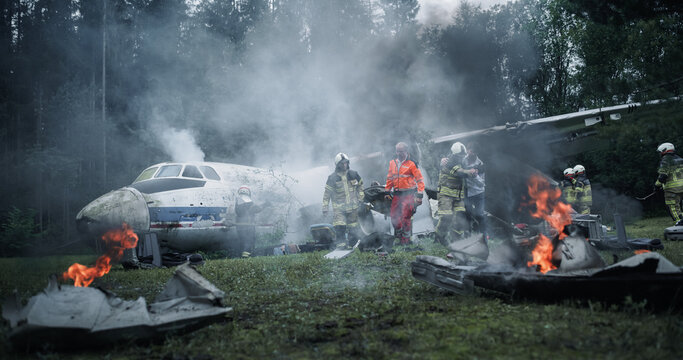 Airplane Rescue Firefighters and EMS Staff in Orange High-visibility Gear Work Beside a Crashed Plane Amid Smoke, Flames, Scattered Wreckage in a Forest Crash Survivor Rescue Scene.