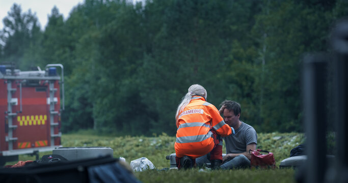 Female Paramedic Kneels on Grass to Assess an Injured Male Survivor After an Airplane Crash, Giving First Aid Beside Fire Truck Stands by During the Active Aviation Emergency Response.