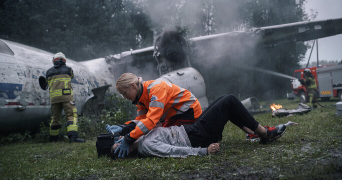 Female Paramedic Supports an Injured Survivor on the Grass Beside a Wrecked Airplane as Firefighters Cool the Smoking Engine and Fuselage During an Ongoing Coordinated Crash Response Operation.