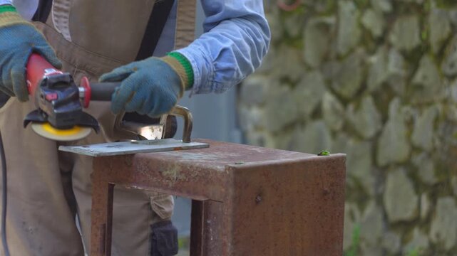 A close-up view of a person using an angle grinder to cut a metal plate securely clamped to a weathered, rusty industrial workbench outdoors.