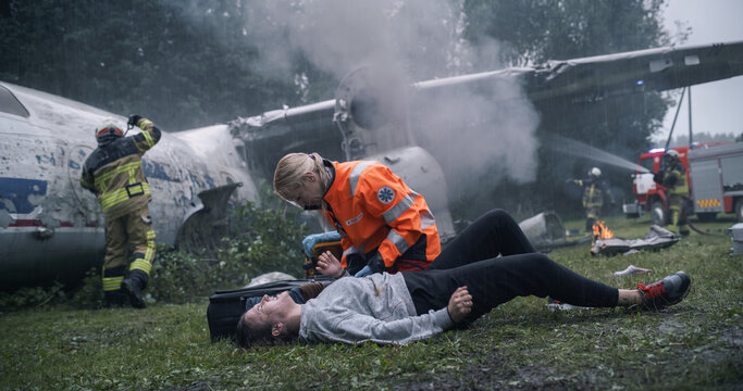 Paramedic Checks an Injured Woman on the Ground as Firefighters Spray Water on a Crashed Airplane, Working Beside Smoking Wreckage and Debris During an Active Aviation Accident Rescue Operation.