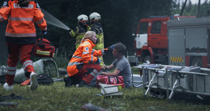 Emergency Crews Respond to an Airplane Crash as a Female Paramedic Treats a Seated Male Survivor on the Ground, While Firefighters Hose Nearby Wreckage and a Fire Engine and Stretchers Support Triage.