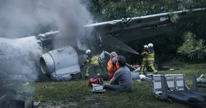 Firefighters Spray Water on a Smoking Airplane Wreck While a Female Paramedic Treats a Seated Male Survivor on the Grass, With Rescue Gear, Detached Seats and Debris Scattered Around the Crash Scene.