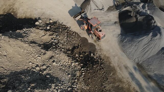 Drone view of a wheel loader transporting a large boulder across an active quarry mining site. The aerial perspective highlights heavy machinery, raw materials, and earthmoving operations 