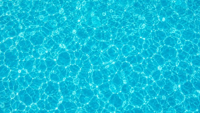 A top-down aerial photograph of a crystal-clear swimming pool surface featuring gentle ripples and shimmering caustic light patterns.