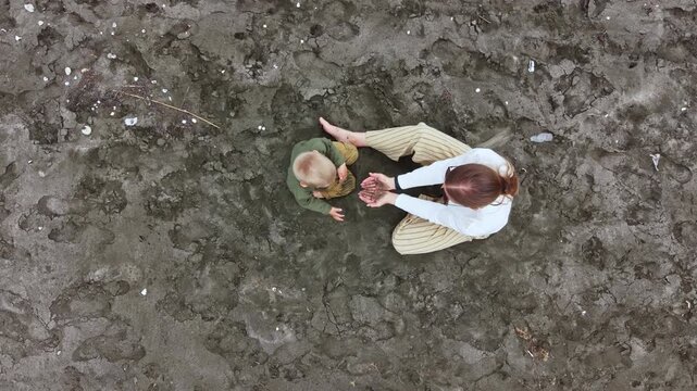 A mother sits in wet sand with her child at the beach during low tide. They play together while surrounded by footprints and patches of water in the late afternoon light.