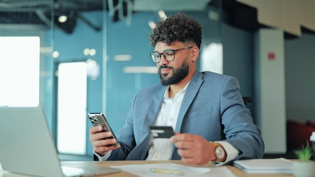 Focused Middle Eastern businessman in glasses holding a credit card and smartphone to make an online payment at his office desk. Corporate executive shopping or verifying banking transactions.