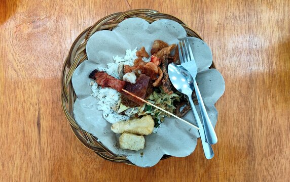 Balinese nasi babi guling with rice, pork meat, pork satay, crispy pork skin and vegetables served on paper in a woven basket.