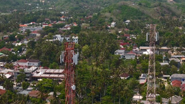 Aerial view shows hillside tropical town with telecom towers scattered houses palm trees and roads for island infrastructure local community and travel documentary projects