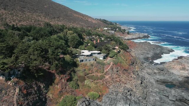 Fly-through over rugged clifftop with modern villas integrated into dense native vegetation above dramatic rocky coastline with crashing waves. Punta Pite, Papudo, Chile