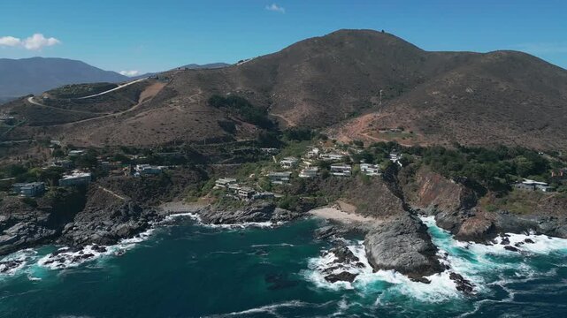 Sliding shot along dramatic rocky coastline with turquoise crashing waves, exclusive clifftop villas and dry scrubland hills rising behind. Punta Pite, Papudo, Chile